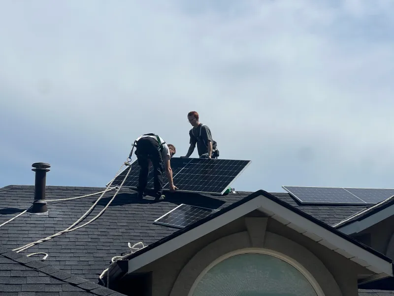 Two installers setting a panel on a shingle roof under a cloudy sky